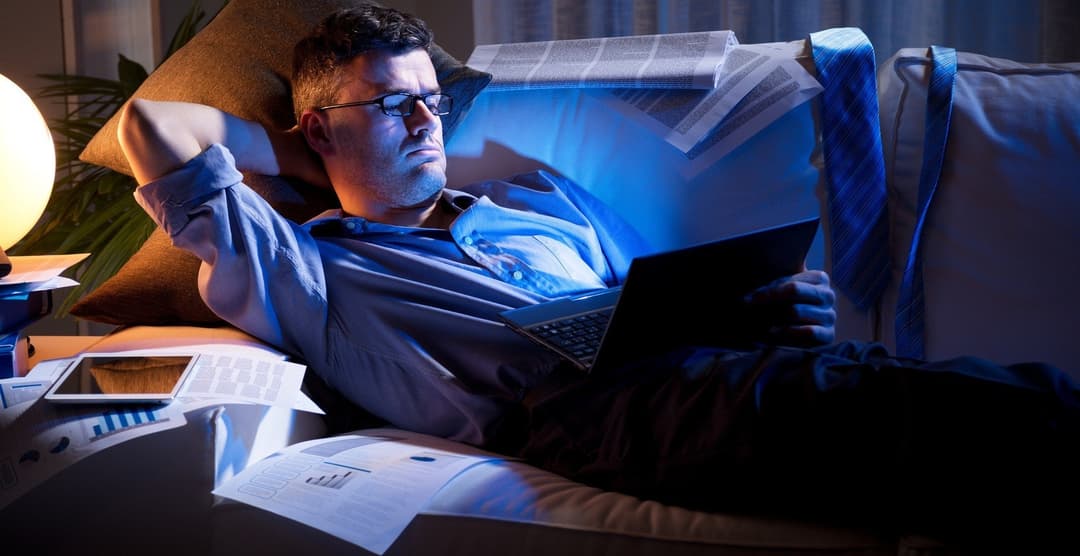 Businessman lying down on sofa and working with laptop late at night.
