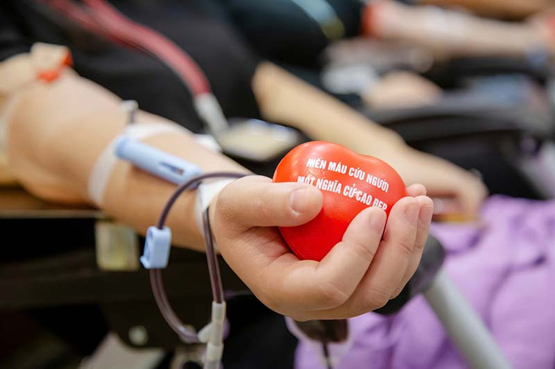 Người đang hiến máu cầm bóng bóp màu đỏ, tay được nối dây truyền máu.